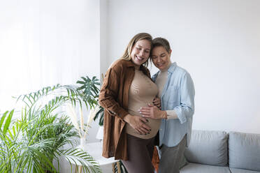 Smiling mother touching belly of pregnant daughter standing at home - AAZF00405