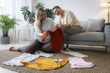 Mother and pregnant daughter looking at baby clothes sitting in living room at home - AAZF00412