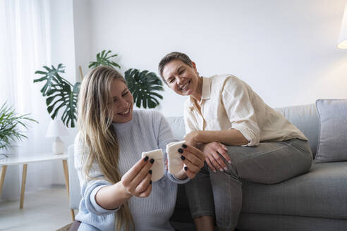 Schwangere Tochter zeigt der Mutter auf dem Sofa sitzend Babysocken - AAZF00413