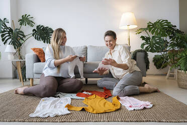 Happy mother and pregnant daughter looking at baby clothes sitting at home - AAZF00415