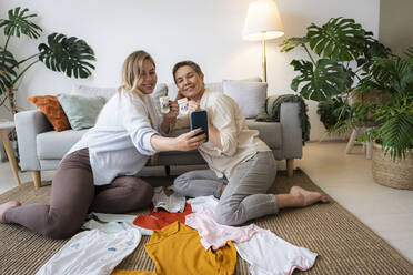 Pregnant daughter taking selfie with mother holding baby socks sitting at home - AAZF00417