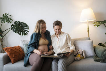 Mutter und schwangere Tochter teilen sich ein Fotoalbum auf dem Sofa zu Hause - AAZF00418