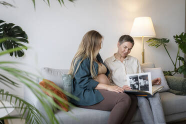 Mutter schaut sich ein Fotoalbum an, während sie mit ihrer schwangeren Tochter zu Hause sitzt - AAZF00420