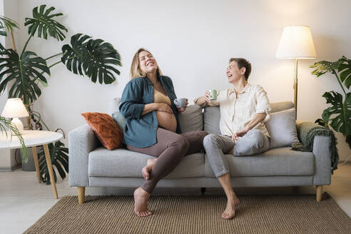 Glückliche schwangere Tochter, die mit ihrer Mutter auf dem Sofa zu Hause Kaffee trinkt - AAZF00423