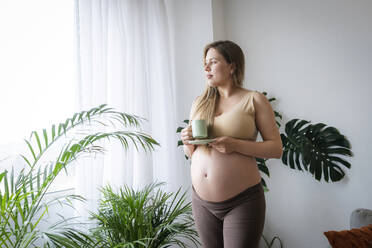 Pregnant woman having coffee standing near plants at home - AAZF00435