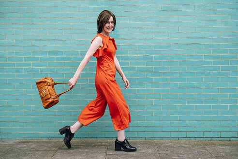 Smiling woman wearing jumpsuit walking by brick wall - AMWF01314