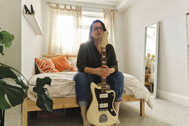 Woman sitting with guitar on bed at home - ASGF03586