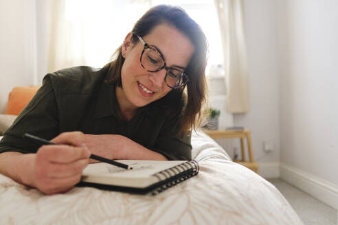 Smiling woman sketching with pencil in bedroom at home - ASGF03588