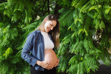 Lächelnde schwangere Frau mit Händen auf dem Bauch vor einem Baum stehend - DCRF01575