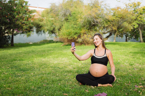 Glückliche schwangere Frau nimmt Selfie sitzen im Schneidersitz auf Gras - DCRF01595
