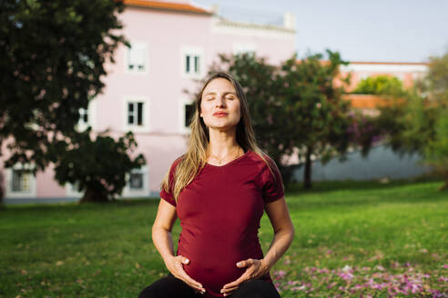 Schwangere Frau meditiert mit Händen auf dem Bauch auf dem Rasen - DCRF01604