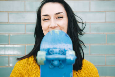 Glückliche junge Frau mit schwarzen Haaren hält blaues Skateboard vor einer Wand - AMWF01327