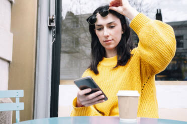 Frau in gelbem Pullover hebt Sonnenbrille und benutzt Smartphone in einem Café im Freien - AMWF01334