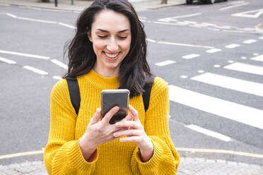 Smiling female tourist in yellow sweater using smartphone near city street - AMWF01338
