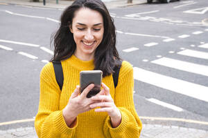 Lächelnde Touristin im gelben Pullover benutzt ihr Smartphone in der Nähe einer Straße - AMWF01338