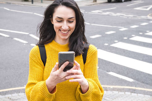 Lächelnde Touristin im gelben Pullover benutzt ihr Smartphone in der Nähe einer Straße - AMWF01338
