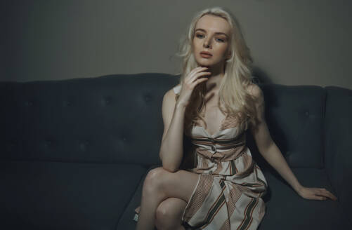 Blonde Frau mit Hand am Kinn sitzt auf Sofa in Dunkelkammer - AZF00518