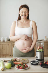 Lächelnde schwangere Frau mit Händen auf dem Bauch, die zu Hause steht - ALKF00280