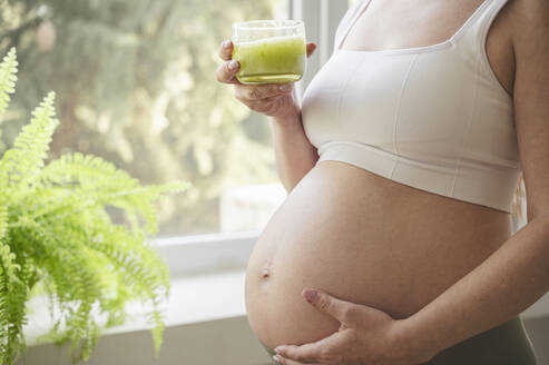 Schwangere Frau mit Hand auf dem Bauch, die ein Glas grünen Smoothie zu Hause hält - ALKF00281