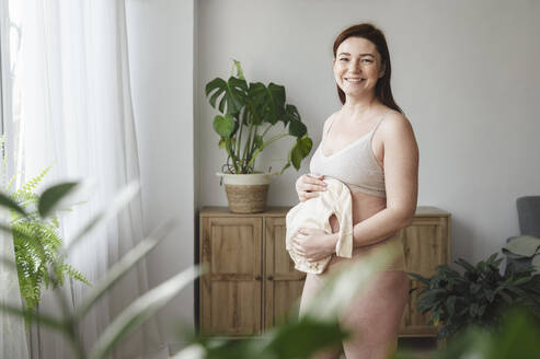 Lächelnde schwangere Frau mit Babykleidung zu Hause - ALKF00283