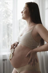 Schwangere Frau mit Hand auf der Hüfte hält Babysocken zu Hause - ALKF00285