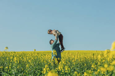 Vater hebt Sohn inmitten von Blumen in einem Rapsfeld hoch - VSNF00772