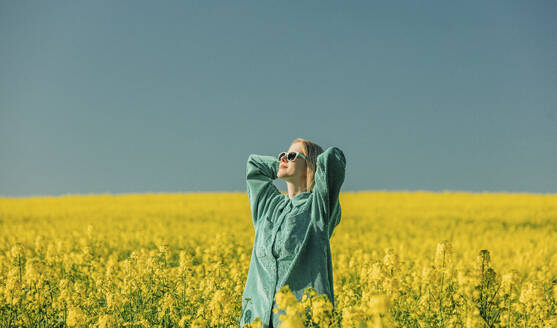 Woman with hands behind head standing in rapeseed field - VSNF00777