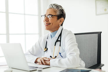 Erfahrene Ärztin, die in ihrem Büro vor einem Laptop sitzt und nachdenklich lächelt. Ältere Ärztin, die in ihrem Büro arbeitet und einen Laborkittel und ein Stethoskop um den Hals trägt. - JLPSF29557