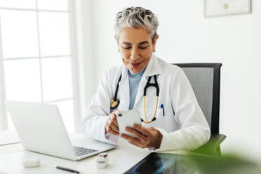 Mature female doctor using a smartphone in her office. The experienced, gray-haired physician is making use of modern technology in her medical practice, easily and efficiently providing healthcare to her patients. - JLPSF29559