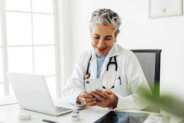 Mature female physician having a video call with a patient on a smartphone, smiling as she hears news of recovery during the virtual consultation. Experienced medical doctor practicing telemedicine in her office. - JLPSF29560