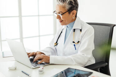 Ältere Ärztin, die einen Laptop benutzt, um auf medizinische Software zuzugreifen, mit der sie effizient praktizieren kann. Ärztin, die in ihrem Büro mit Technologie arbeitet, trägt einen weißen Laborkittel und ein Stethoskop am Hals. - JLPSF29566