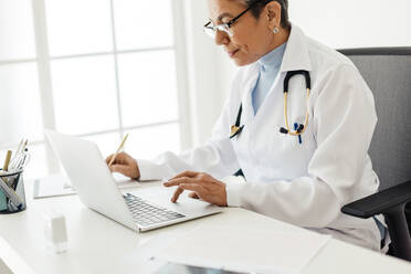 Mature medical doctor using EHR software to access patient records in her office, copying information from her laptop to a clipboard. Experienced female physician writing a prescription in her office. - JLPSF29567
