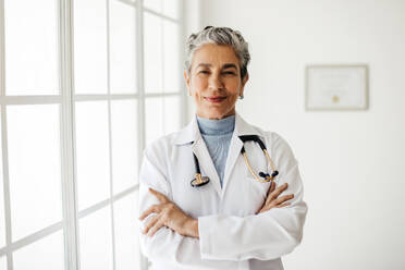 Porträt einer reifen Ärztin im weißen Kittel, die mit verschränkten Armen in ihrem Büro steht und ihr Vertrauen in ihre Praxis als erfahrene Medizinerin ausdrückt. - JLPSF29570