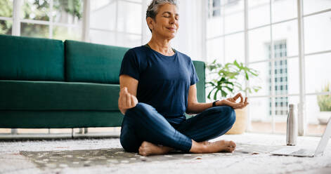 Gesunde ältere Frau meditiert im Lotussitz, während sie an einem Online-Fitnesskurs teilnimmt. Ältere Frau, die zu Hause vor dem Laptop Yoga praktiziert und im Ruhestand Entspannung und Achtsamkeit findet. - JLPSF29573