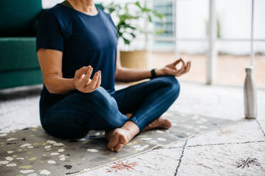 Eine ältere Frau macht eine entspannende Atemübung, während sie zu Hause Yoga-Meditation praktiziert. Auf einer Fitnessmatte im Lotussitz sitzend, erlangt sie Achtsamkeit und inneren Frieden durch diese Trainingsroutine. - JLPSF29575