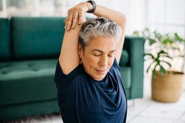 Ältere Frau bei einer Armdehnung für einen gesunden Trizeps in einer ruhigen Workout-Sitzung zu Hause. Aktive Frau mit grauem Haar, die Yoga für ein besseres körperliches Wohlbefinden im Rentenalter praktiziert. - JLPSF29579