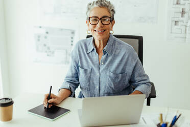 Successful female designer smiling at the camera while drawing with a graphics tablet in her office. Senior graphic designer using her expertise and advanced tech tools to create excellent designs. - JLPSF29584