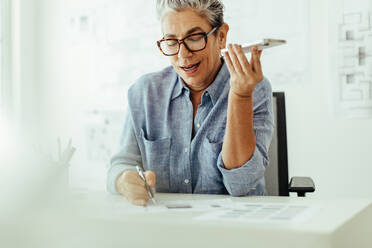 Creative business woman discussing a proposal with a client on a phone call, using a document to make confirmation on the details. Senior interior designer planning another client project in her office. - JLPSF29586