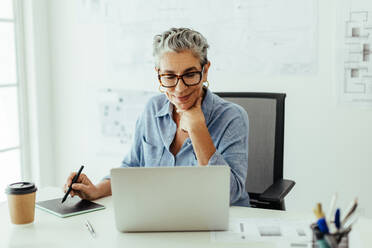 With years of experience in design, this senior graphic designer creates stunning visuals at her desk using a laptop and graphics tablet. Senior woman looking pleased with her creative work in an office. - JLPSF29588