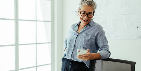 Ältere Architektin, die in einem Büro steht und mit einem Lächeln im Gesicht eine Textnachricht liest. Kreative Geschäftsfrau, die ein Smartphone benutzt, um mit ihren Kunden in der Designbranche in Kontakt zu treten und zu kommunizieren. - JLPSF29594