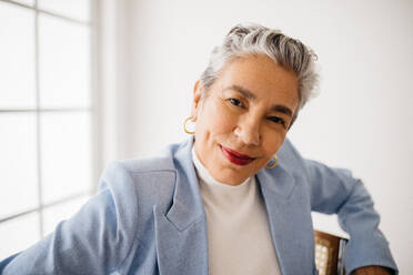 Woman with greying hair looking at the camera with confidence and self-assurance in her expertise as a business professional. Mature woman sitting in an office, dressed in an elegant business attire. - JLPSF29602