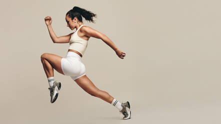 Sportswoman jumping forward, running in a vigorous studio workout. Wearing sportswear, she combines strength training with cardio exercises to improve fitness and performance. Female athlete pushing herself to new limits. - JLPSF29625