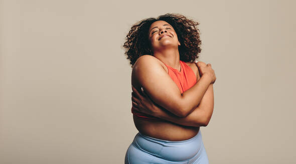 Body confident woman embracing her body, expressing self-love and self-acceptance. Young plus-size woman standing in a studio in fitness clothing, embracing her natural physique with joy. - JLPSF29695