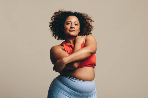 Body positive woman looking at the camera as she confidently embraces her body, expressing self-love. Plus-size woman with curves and an athletic physique stands in a studio wearing fitness clothing. - JLPSF29696