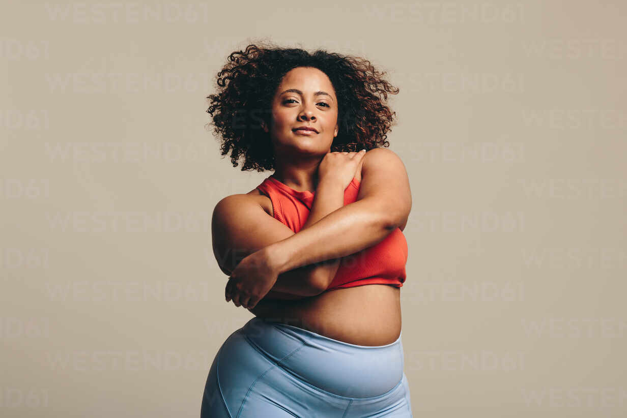Body positive woman looking at the camera as she confidently embraces her  body, expressing self-love. Plus-size woman with curves and an athletic  physique stands in a studio wearing fitness clothing. stock photo