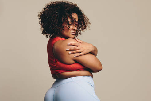 Fit young woman confidently embracing her body in a sportswear outfit, radiating body positivity and self-love. Plus size woman with an athletic physique and a curly afro standing in a studio. - JLPSF29697