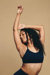 Eine junge erwachsene Frau steht mit erhobenen Armen im Studio, um ihre Stärke und Vitalität zu demonstrieren. Sie trägt Sportkleidung und eine modische braune Frisur und verkörpert Positivität durch körperliche Fitness. - JLPSF29700