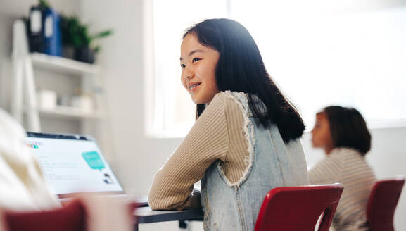 Young Asian girl sitting in a coding class with a laptop in front of her and a keen interest on her face. Eager to master her programming skills, she pays attention to the lesson in the positive learning environment. - JLPSF29704