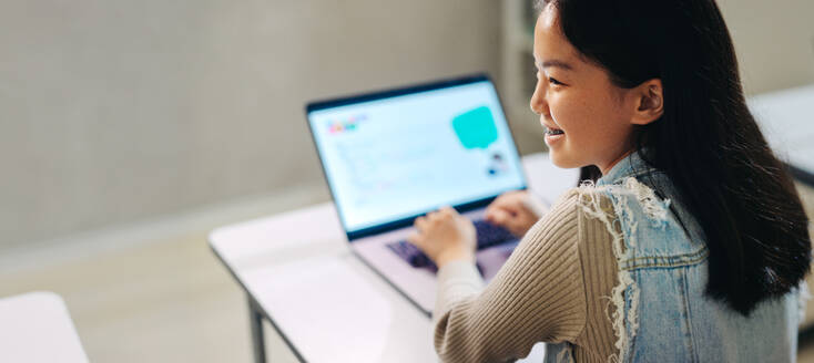 Happy Asian girl learning how to code on a laptop. Sitting in a computer literacy classroom, she engages in the lesson and takes part in a programming exercise. Inspired female child developing her tech skills in a modern learning environment. - JLPSF29706