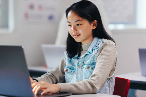 Asian kid nurturing her newly discovered tech talent as she learns to code on a laptop. Sitting in a computer science classroom, the young girl takes on a programming exercise given to the class, mastering her digital literacy skills. - JLPSF29708